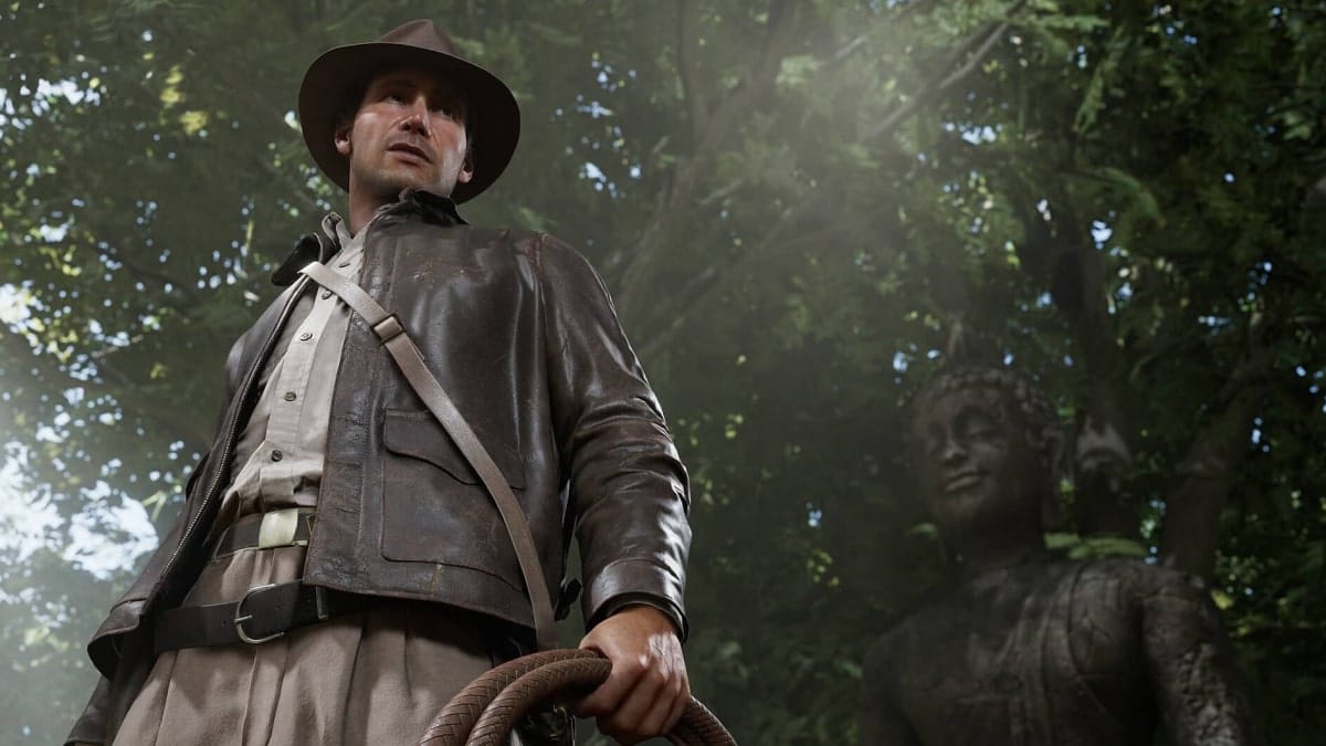 Indiana Jones stands in a jungle clearing holding his whip beside an ancient statue